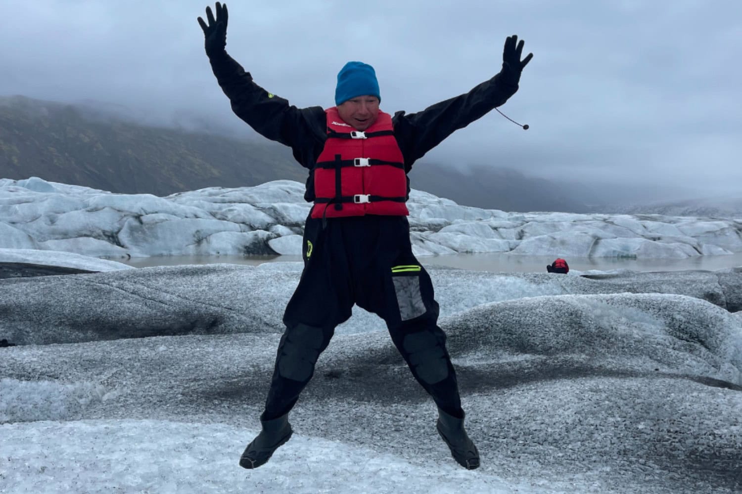Mężczyzna w skafandrze i kamizelce asekuracyjnej skacze na lodowcu. W tle widać zlodowaciały krajobraz i pochmurne niebo.