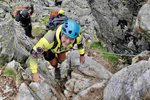 Mężczyzna w kapeluszu i stroju trekkingowym stoi na drewnianym moście z kijkami do chodzenia. W tle widać gęstą roślinność i górski szczyt w chmurach.