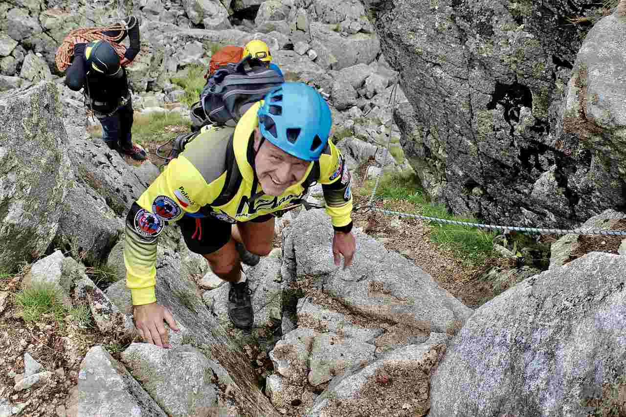 Mężczyzna w kapeluszu i stroju trekkingowym stoi na drewnianym moście z kijkami do chodzenia. W tle widać gęstą roślinność i górski szczyt w chmurach.