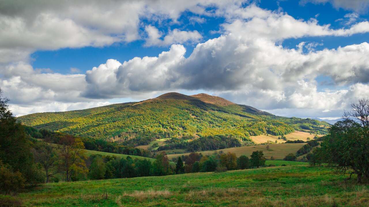 Panorama gór pokrytych lasami i łąkami pod częściowo pochmurnym niebem. Na pierwszym planie zielone pastwiska.
