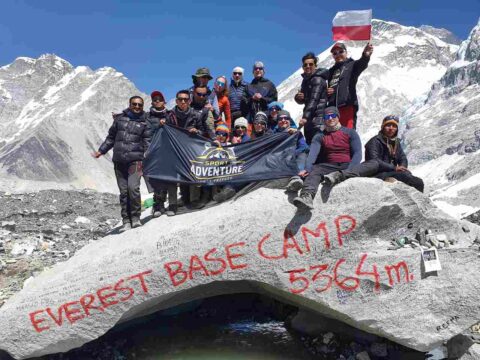 Grupa uczestników wyprawy pozuje na kamieniu z napisem „Everest Base Camp 5364 m”. Trzymają flagę Sport Adventure i flagę Polski. W tle widać ośnieżone szczyty Himalajów.
