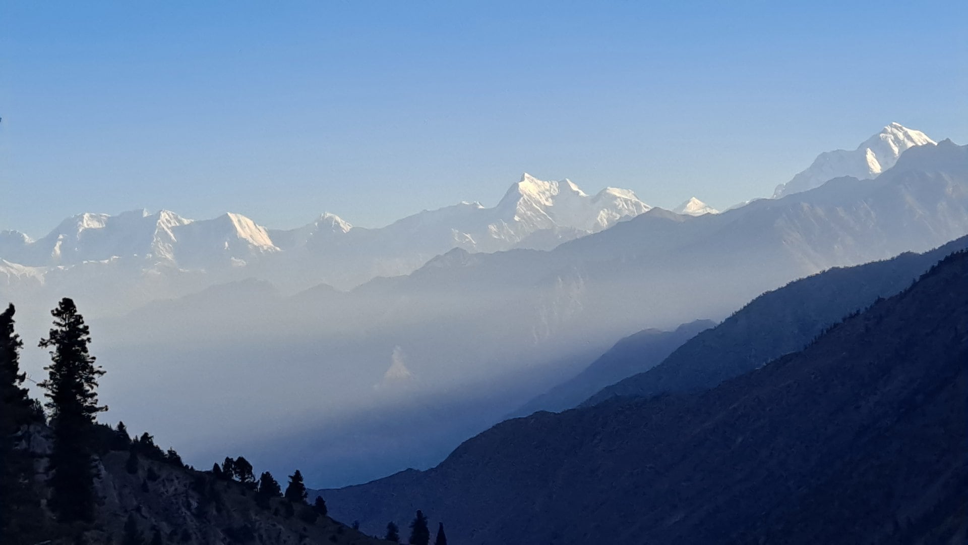 Panorama ośnieżonych szczytów górskich o wschodzie słońca. Na pierwszym planie ciemne zbocza i drzewa, w oddali widać oświetlone przez słońce wierzchołki gór i warstwę porannej mgły w dolinie.