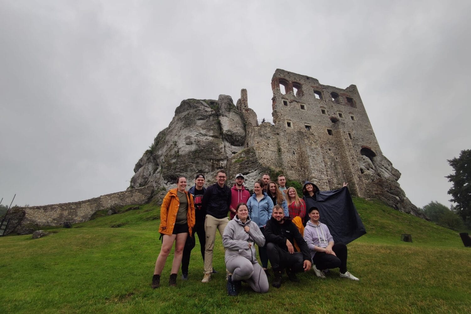 Grupa osób pozuje do wspólnego zdjęcia na tle ruin kamiennego zamku stojącego na skale.