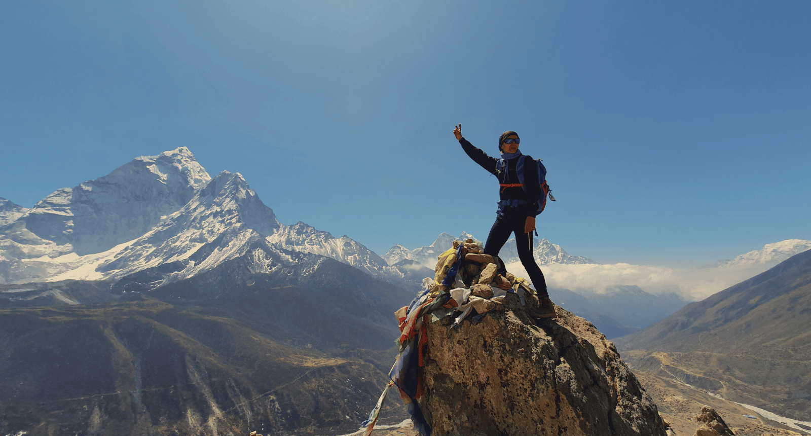 Osoba stojąca na skalnym szczycie w Himalajach, unosząca rękę. W tle ośnieżone szczyty, w tym Ama Dablam.