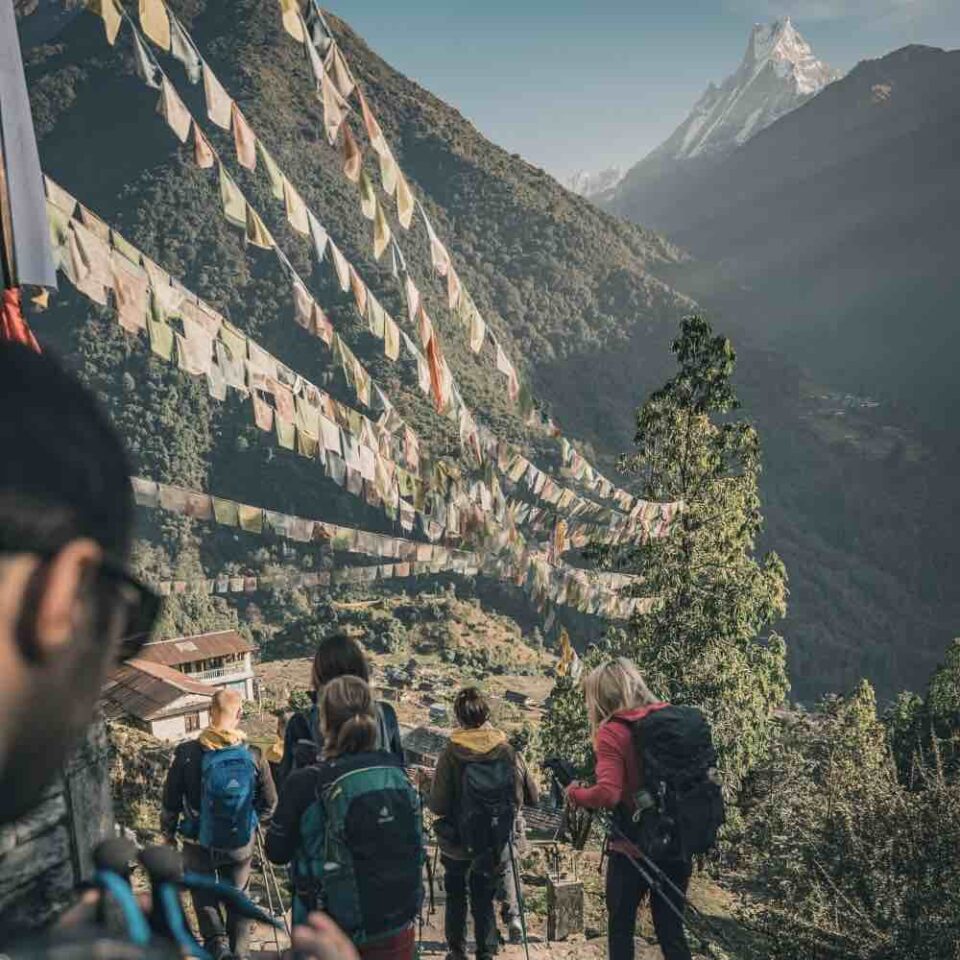 Trekking w Nepalu – grupa turystów schodząca górskim szlakiem z widokiem na Machapuchare i buddyjskie flagi modlitewne rozwieszone nad doliną Annapurny.