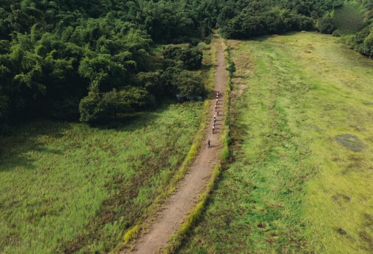 Widok z lotu ptaka na długą, piaszczystą ścieżkę biegnącą przez zielone pola, z grupą rowerzystów jadących w szeregu, obok gęstego lasu w Mũi Né, Wietnam.