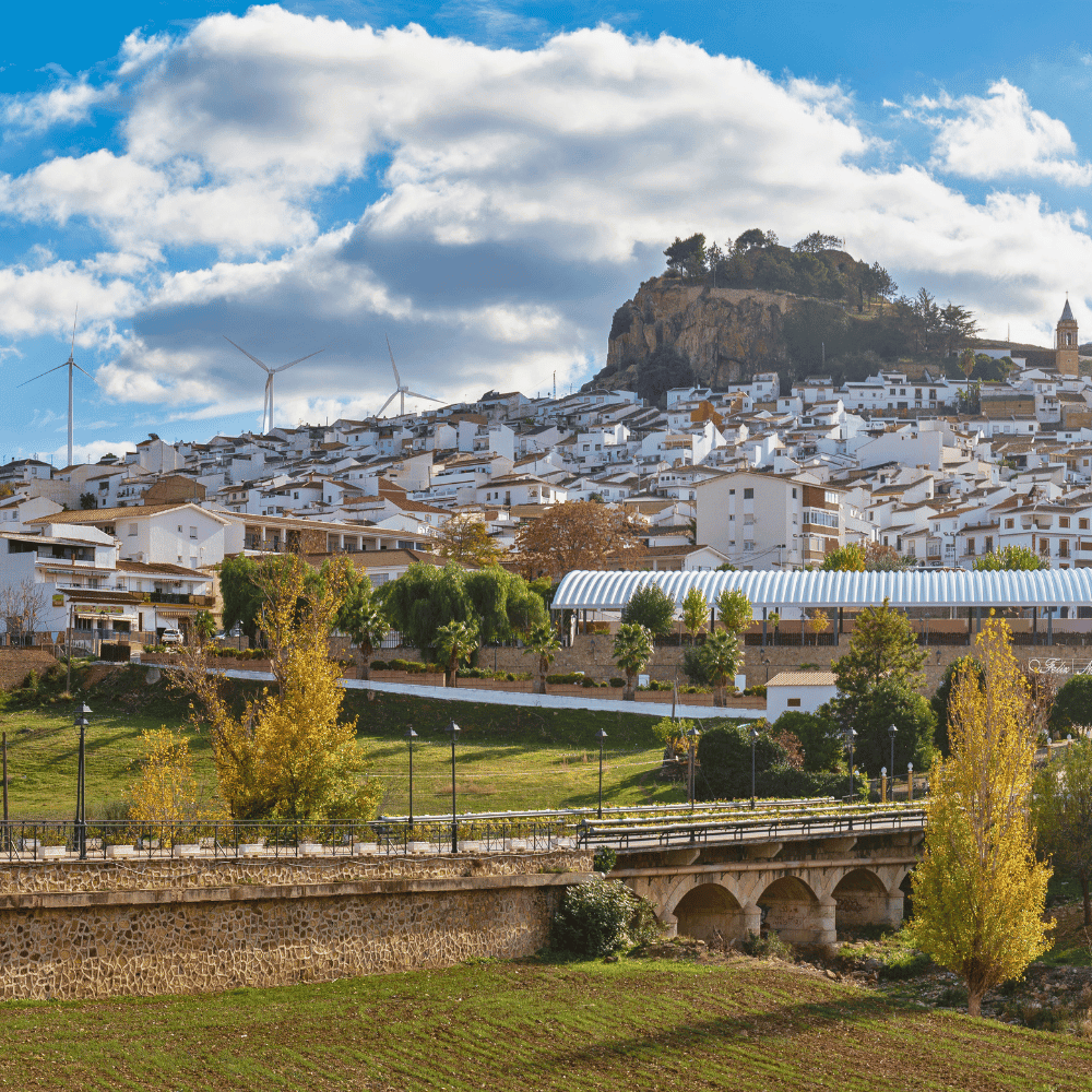Panoramiczny widok białego miasteczka Álora rozłożonego na zboczu wzgórza, za którym na horyzoncie stoją wiatraki elektrowni wiatrowej, a nad tym wszystkim płyną obłoki na niebie.