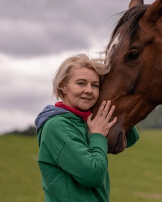 Kobieta w zielonej bluzie przytula brązowego konia na tle bieszczadzkiego krajobrazu