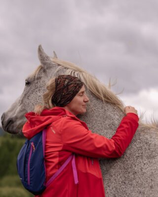 Kobieta w czerwonej kurtce przytula siwego konia podczas wyprawy konnej w Bieszczadach