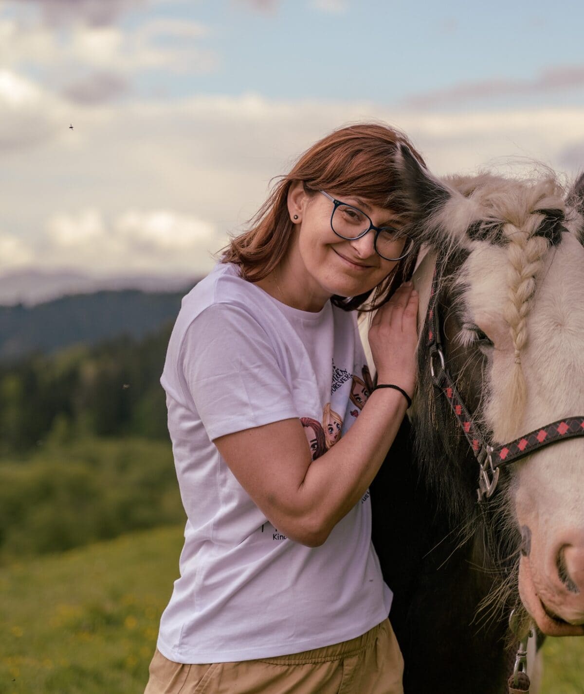 Kobieta w okularach przytula srokatego konia z warkoczem na tle bieszczadzkiego krajobrazu, uśmiechnięta i zrelaksowana.