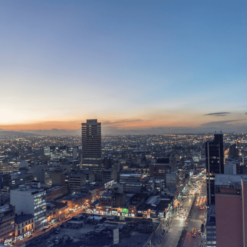 Panorama Bogoty o zmierzchu, z widocznym oświetleniem ulic i sylwetkami wieżowców, na tle pastelowego nieba.