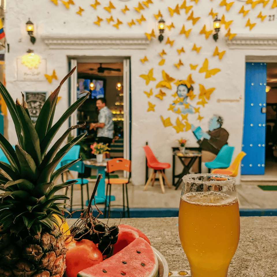 Kawiarnia w Cartagenie z muralem przedstawiającym postać i żółte motyle, z tropikalnymi owocami i szklanką z napojem koloru żółtego.