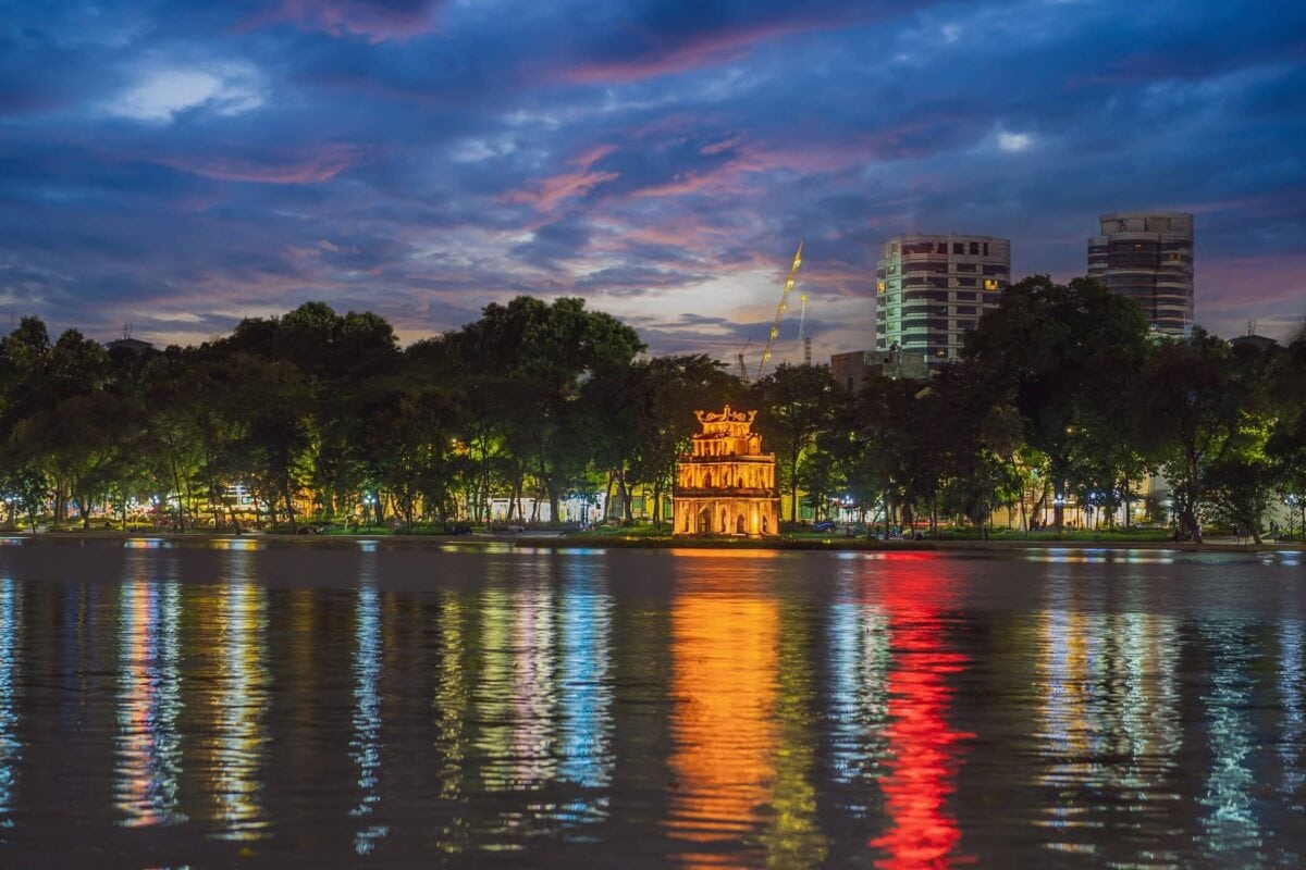 Jezioro Hoan Kiem w Hanoi o zmierzchu z oświetlonym czerwonym mostem The Huc i panoramą miasta w tle.