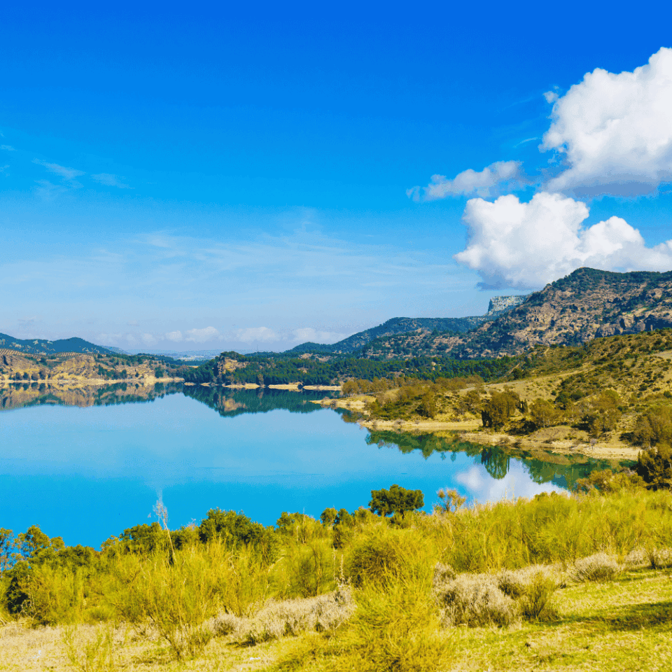Jezioro o intensywnie turkusowej wodzie odbijające wzgórza i chmury, na brzegu żółtozielone zarośla i pasmo pagórków pod błękitnym niebem.