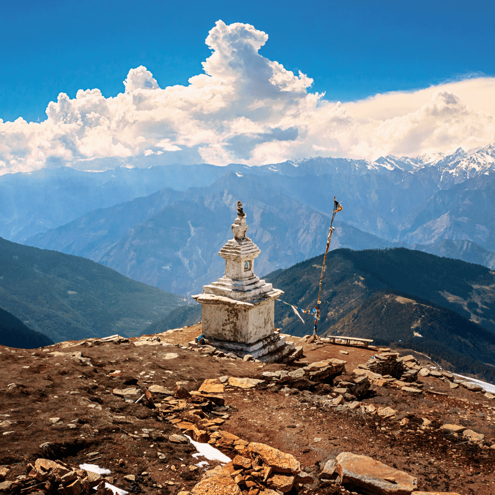 Biała, kamienna stupa stoi na skalistym szczycie góry. W tle rozciągają się rozległe pasma górskie, a nad nimi widać duże, białe chmury na niebieskim niebie.