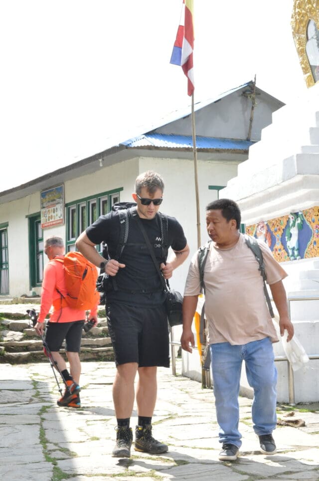 Dwóch mężczyzn idzie razem kamienną uliczką w nepalskiej wiosce; jeden ubrany w czarny strój trekkingowy z plecakiem, drugi w jasnej koszulce i dżinsach, w tle turysta z kijkami i buddyjska stupa z kolorową flagą.