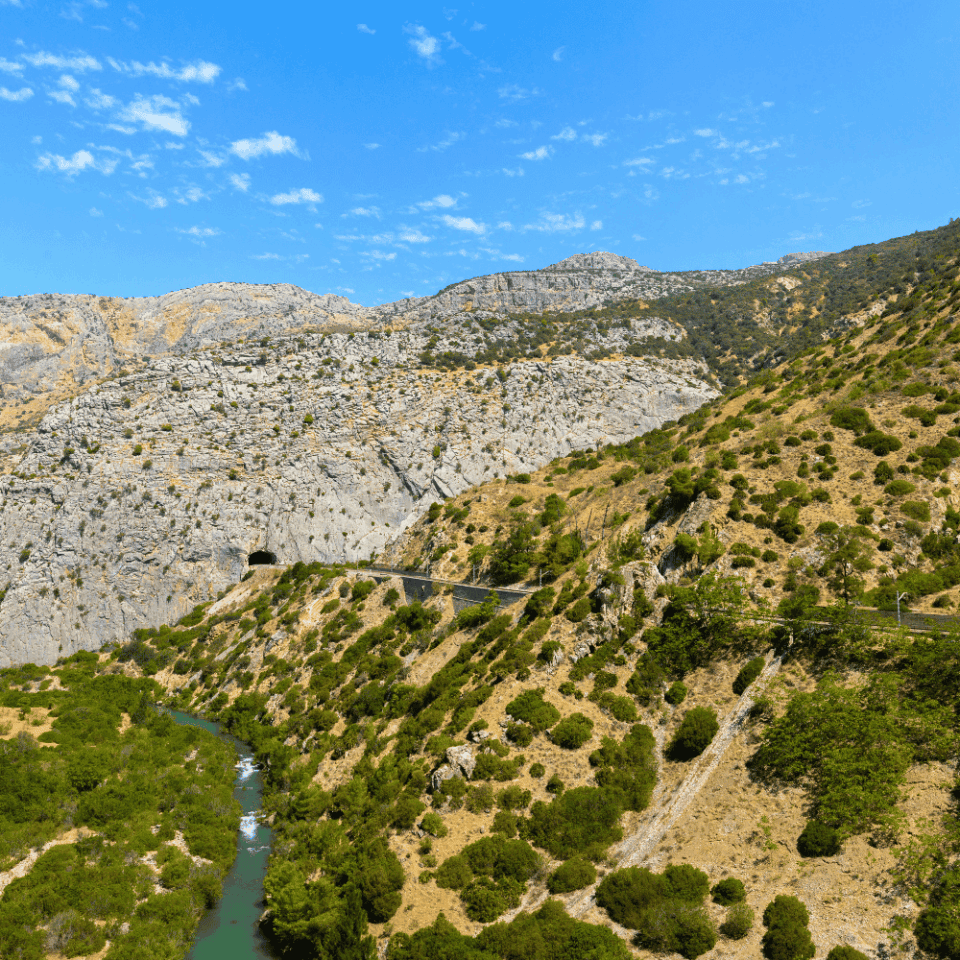 Szeroka perspektywa kanionu El Chorro – w dole wijąca się rzeka, po bokach skaliste urwiska, nad nimi błękitne niebo z kilkoma obłokami i fragmentem drogi kolejowej prowadzącej przez tunele.