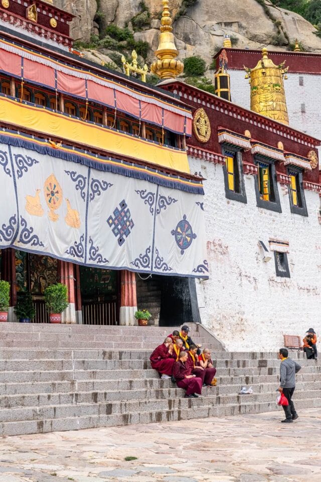 Tybetańskie mniszki w klasztorze Drepung – kobiety w bordowych szatach modlitewnych siedzące na schodach udekorowanych tradycyjnymi wzorami, w tle białe mury klasztoru