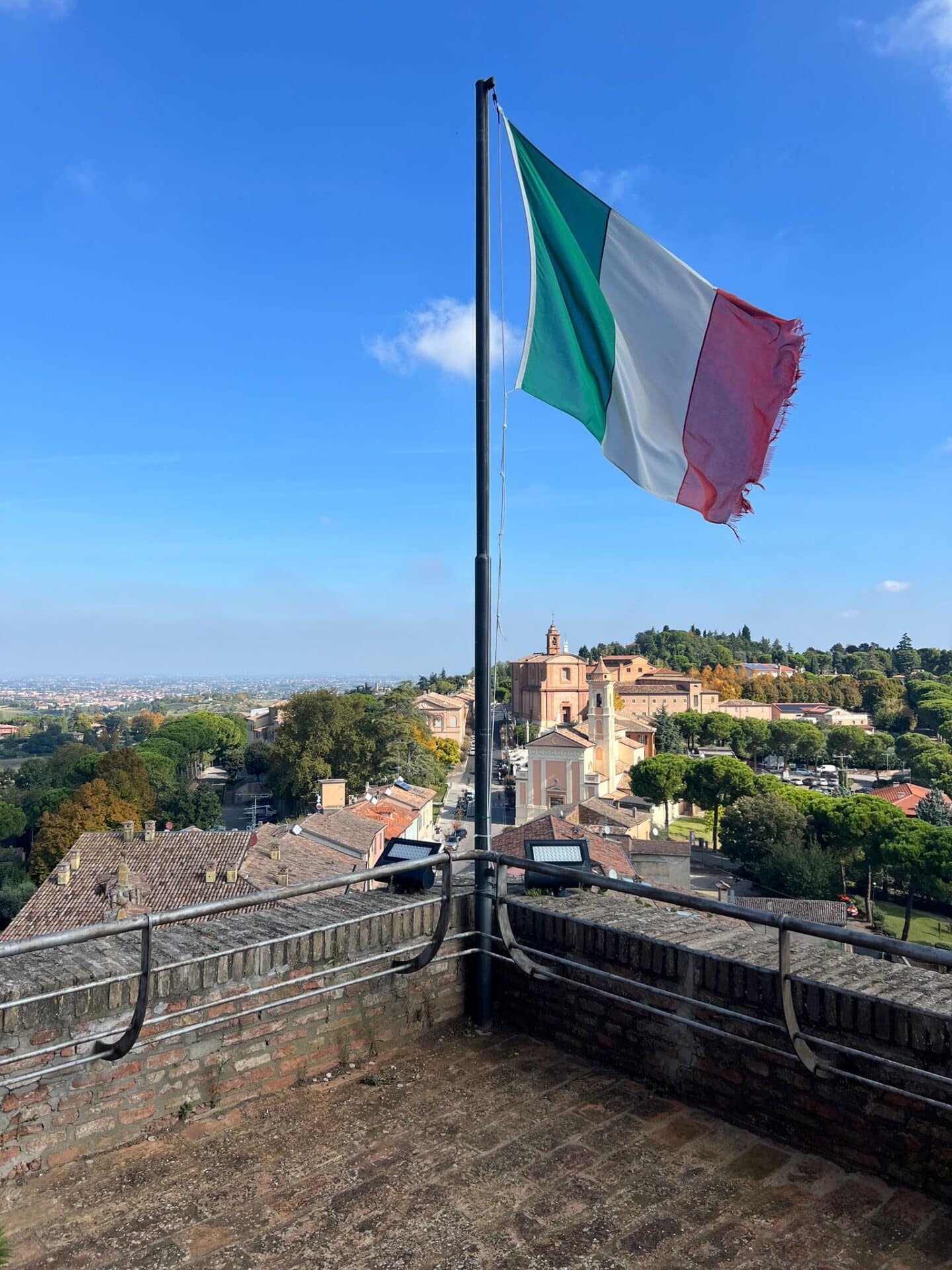 Flaga Włoch powiewająca na wysokim maszcie nad ceglanym murem, w tle malownicze włoskie miasteczko z zabytkową dzwonnicą i zieloną doliną.
