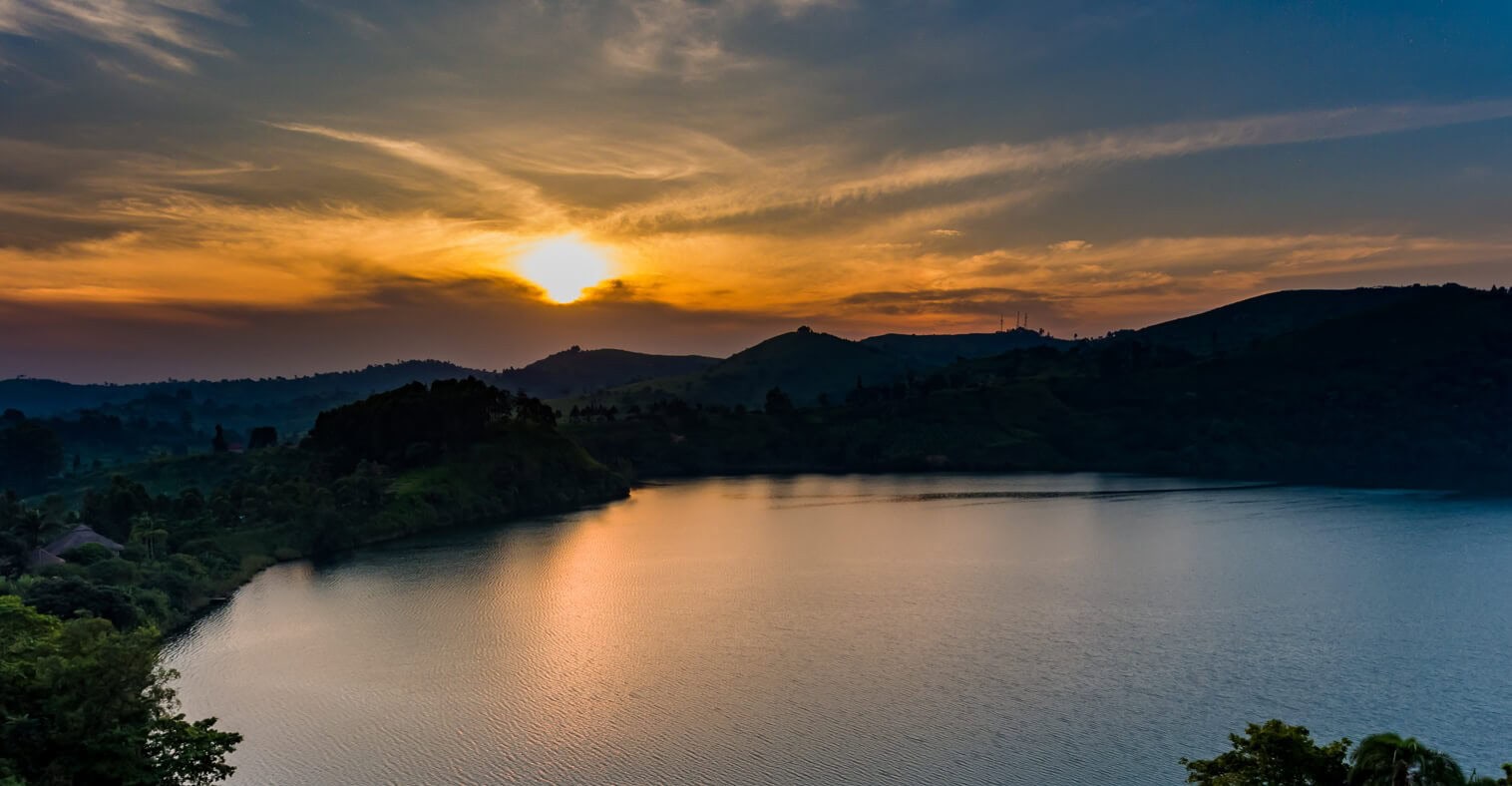 Panorama jeziora kraterowego w Ugandzie podczas zachodu słońca z pomarańczowym blaskiem na niebie i ciemnymi konturami wzgórz.