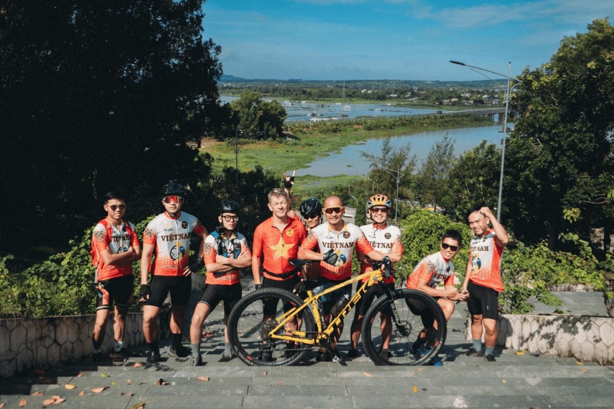 Grupa osób w sportowych strojach kolarskich, pozująca z żółtym rowerem górskim na schodach na tle jeziora i zielonego krajobrazu.