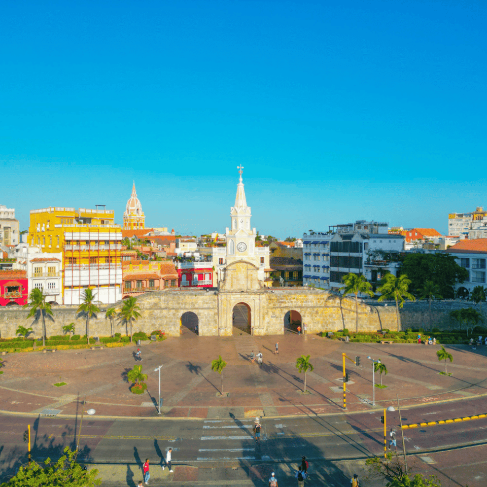 Plac przy Bramie Zegarowej (Torre del Reloj) w Cartagenie, otoczony kolorowymi kolonialnymi budynkami i palmami, przy bezchmurnym niebie.