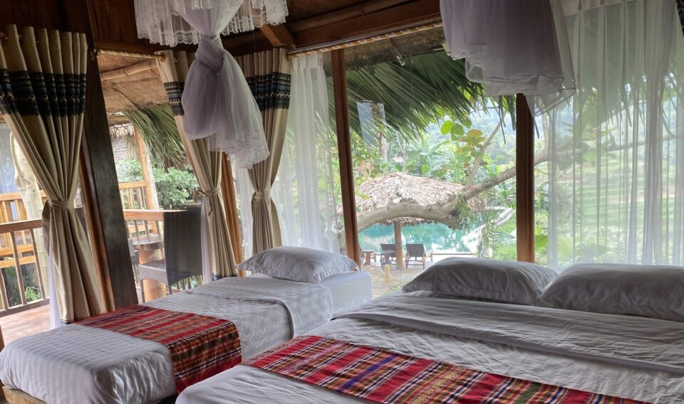 Tradycyjny dom na palach w Wietnamie &ndash; pok&oacute;j z kilkoma ł&oacute;żkami, moskitierami i widokiem na tropikalny ogr&oacute;d.