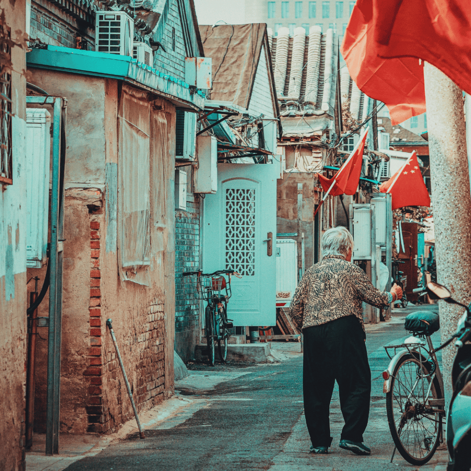 tarsza kobieta przechadza się wąską uliczką hutongu w Pekinie, ozdobioną chińskimi flagami i tradycyjną zabudową.