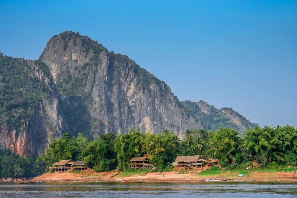 Wejście do jaskiń Pak Ou w Laosie – widok od strony rzeki Mekong, z wapiennym klifem i tropikalną roślinnością u podnóża.
