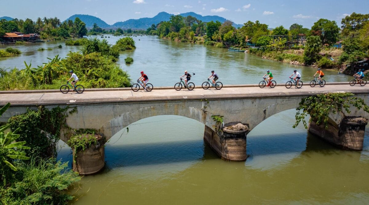 Grupa rowerzystów przejeżdżająca przez most nad rzeką w Laosie.