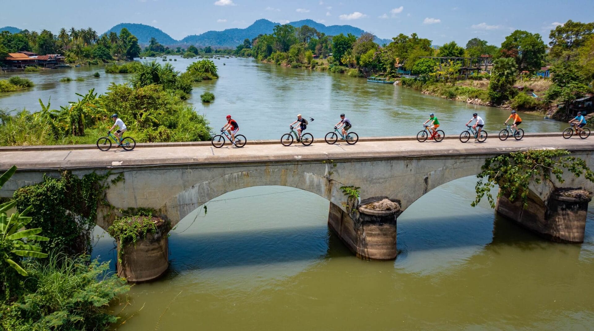 Grupa rowerzystów przejeżdżająca przez most nad rzeką w Laosie.