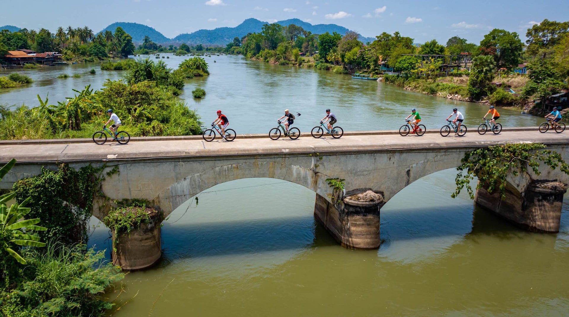 Grupa rowerzystów przejeżdżająca przez most nad rzeką w Laosie.