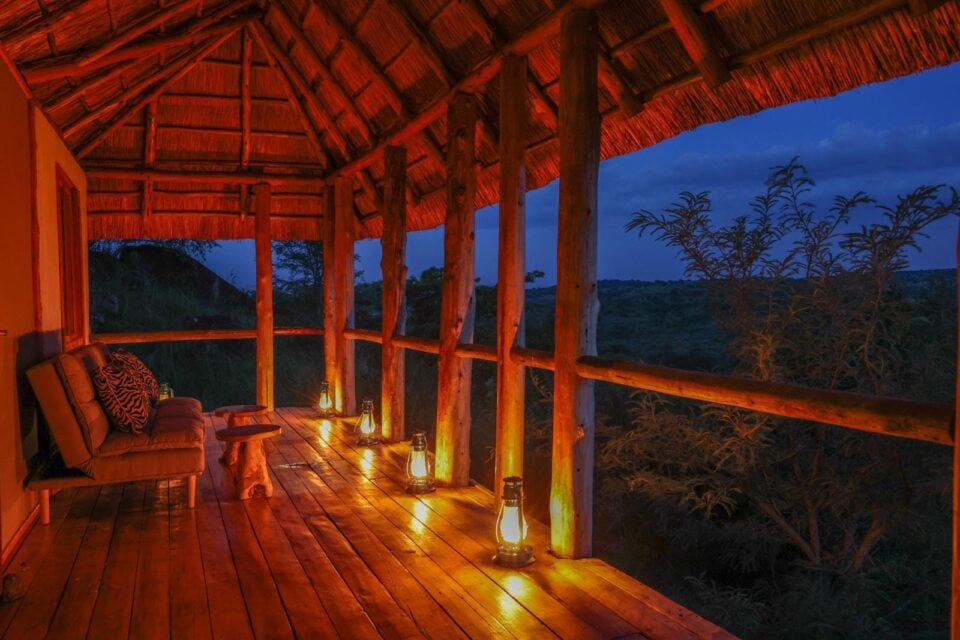 Oświetlony lampionami drewniany taras safari lodge w Ugandzie z kanapą w paski zebry i widokiem na krajobraz sawanny o zmierzchu.