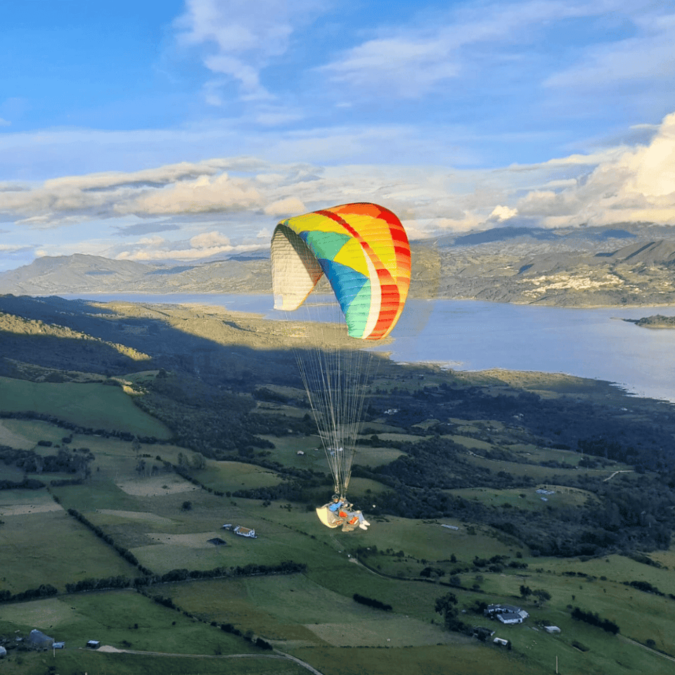 Kolorowy paralotniarz szybujący nad zieloną doliną i rzeką, z widokiem na Kanion Chicamocha i góry w tle.