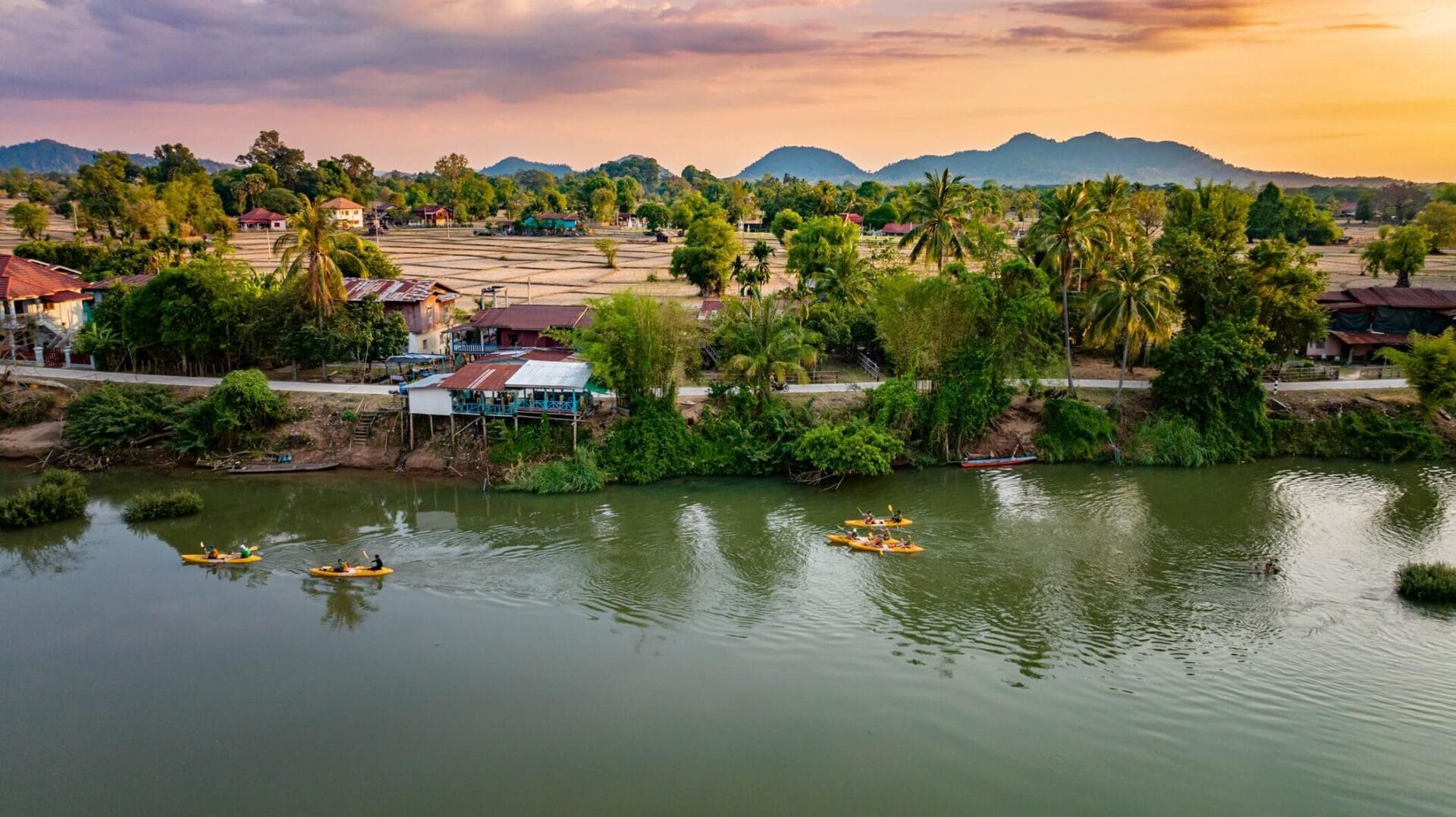Widok na rzekę Mekong i wioskę w regionie Si Phan Don (Cztery Tysiące Wysp) w Laosie – zielone drzewa, tradycyjne domy i kajaki na wodzie o zachodzie słońca.