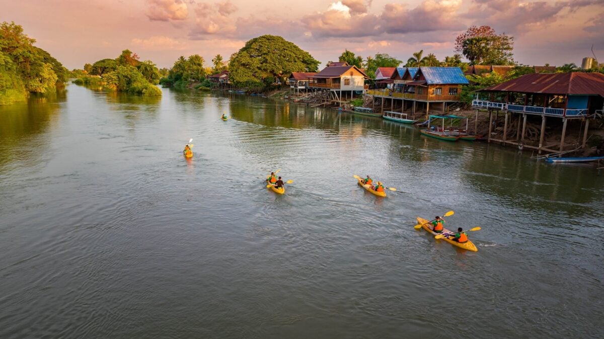 Spływ kajakowy po Mekongu w regionie Si Phan Don (Cztery Tysiące Wysp) w Laosie – o zachodzie słońca, z tradycyjnymi drewnianymi domami na palach w tle.