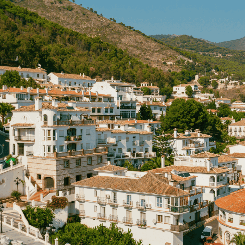 Sielankowy widok na białe domy w miasteczku Mijas z ceglanymi dachami, rozrzucone na zielonym wzgórzu porośniętym sosnami i krzewami, pod jasnym, lekko spowitym chmurami niebem.