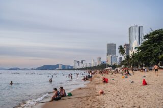 Szeroka piaszczysta plaża z wieloma ludźmi kąpiącymi się i odpoczywającymi, z miejską zabudową i górami w tle, w Nha Trang, Wietnam.