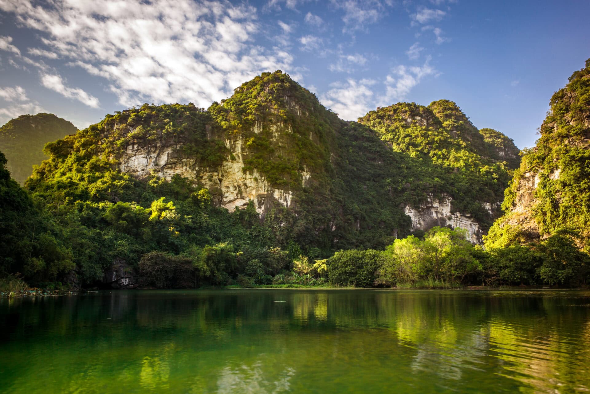 Formacje wapienne w Ninh Binh w Wietnamie, pokryte bujną, tropikalną roślinnością. Klify wznoszą się nad spokojną, szmaragdową wodą, która odbija błękitne niebo z chmurami. U podnóża widoczne wejście do jaskini.