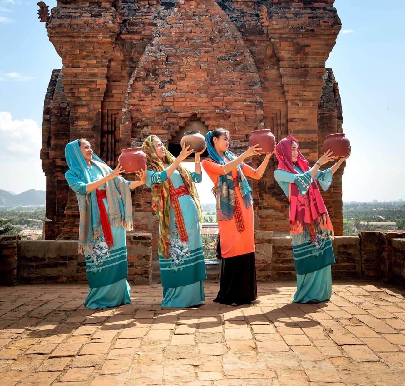 Cztery kobiety w tradycyjnych, kolorowych strojach, trzymające gliniane naczynia, pozują przed starożytną ceglaną świątynią Po Klong Garai w Phan Rang-Tháp Chàm, Wietnam.