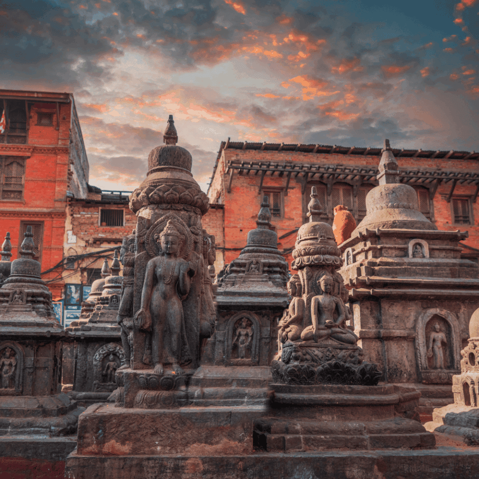 Kamienne rzeźby i stupy na Placu Durbar w Bhaktapur, Nepal, z tradycyjnymi ceglanymi budynkami w tle. Niebo jest częściowo zachmurzone, z pomarańczowo-różowymi odcieniami zachodu słońca.