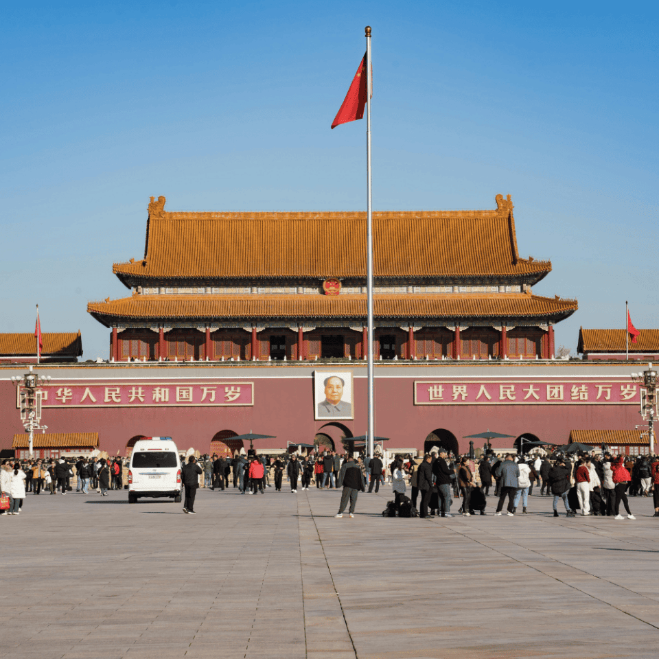 Widok na bramę Tian’anmen z portretem Mao Zedonga i chińską flagą na pierwszym planie, otoczony turystami.