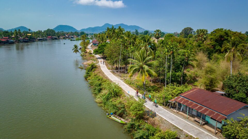 Trasa rowerowa wzdłuż rzeki w Laosie – widok z lotu ptaka na asfaltową drogę otoczoną palmami i wodą.