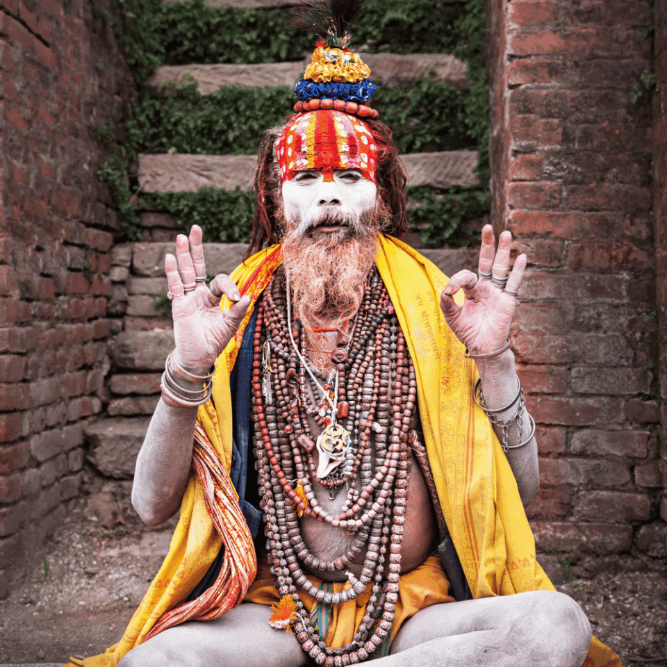 Sadhu (hinduski asceta) siedzący w pozie medytacyjnej na tle ceglanych schodów w Świątyni Pashupatinath w Katmandu, Nepal. Mężczyzna ma pomalowaną twarz na biało, długą, rudą brodę, a jego ciało ozdobione jest licznymi naszyjnikami i turbanem z piórami.