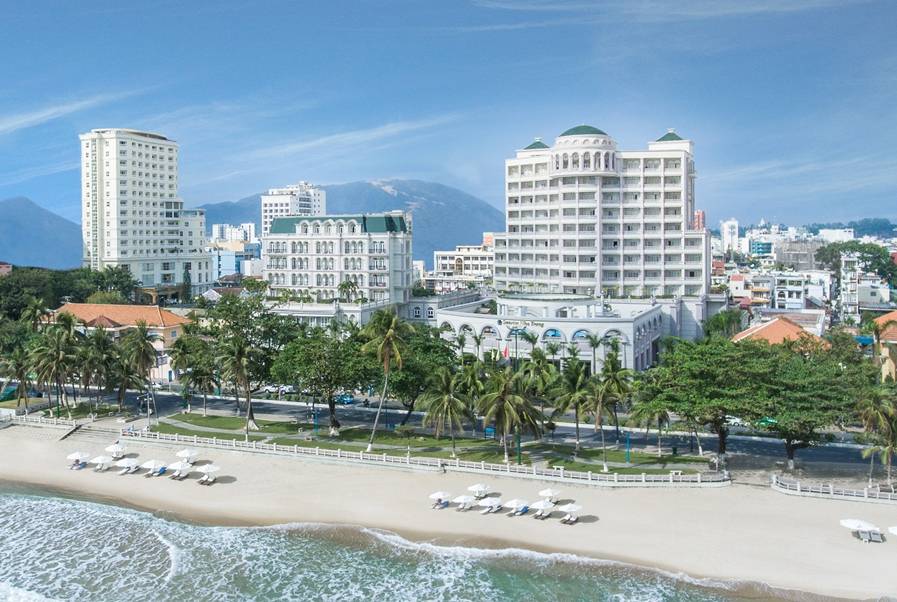 Widok z lotu ptaka na wybrzeże z piaszczystą plażą, rzędem palm i dużymi budynkami hotelowymi, w tym Sunrise Nha Trang Beach Hotel & Spa, z g&oacute;rami w tle, w Nha Trang, Wietnam.