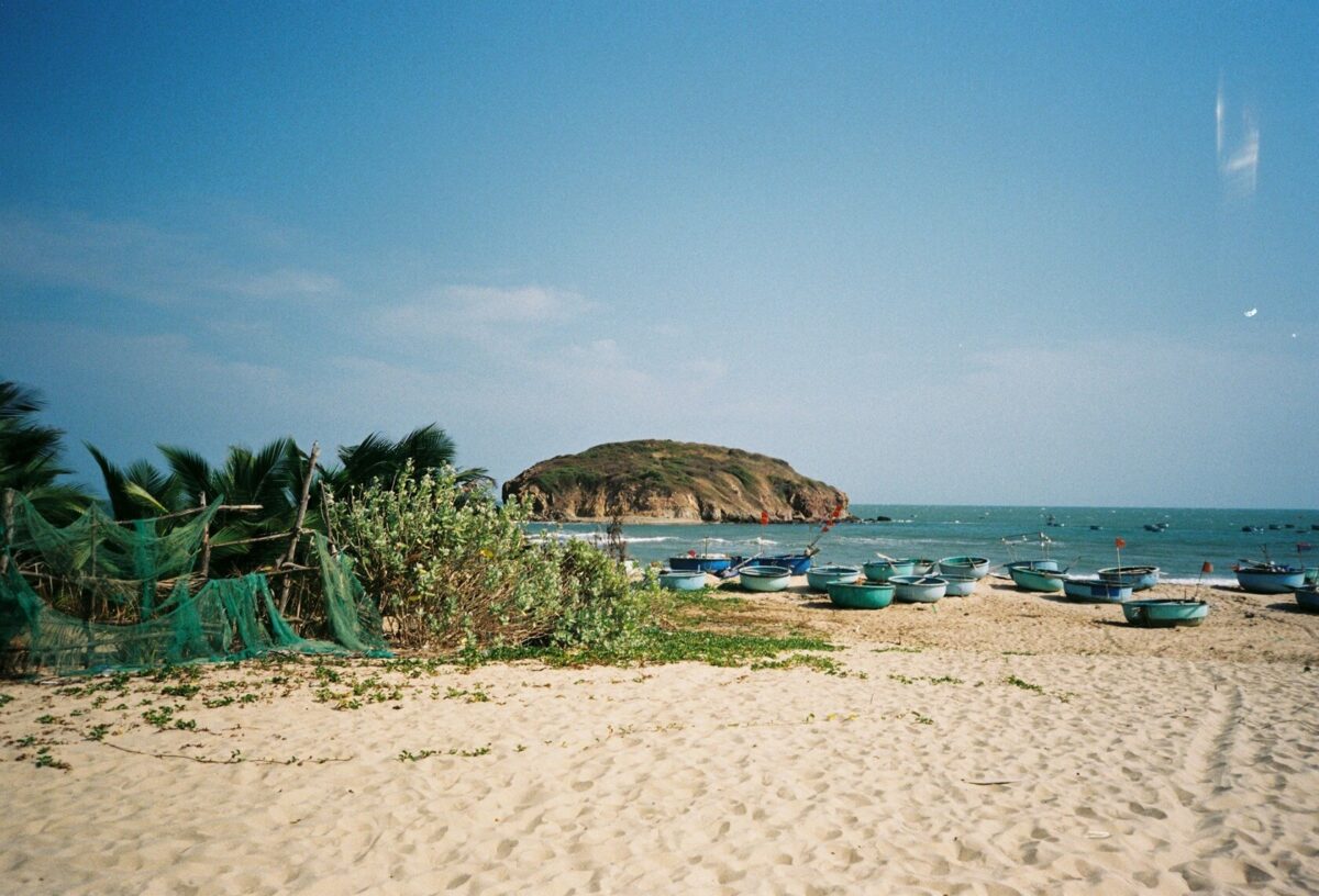 Spokojna piaszczysta plaża z zielonymi krzewami i siatkami rybackimi na pierwszym planie, z licznymi małymi łodziami na wodzie i skalistą wyspą w oddali, pod czystym niebieskim niebem w Mũi Né, Wietnam.