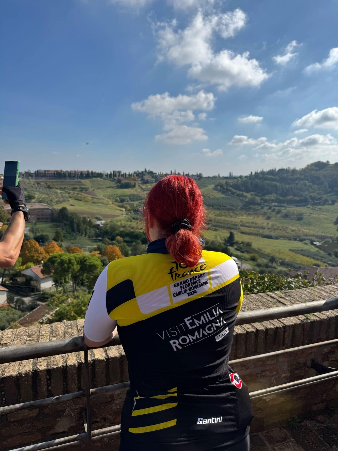 Kobieta o czerwonych włosach w profesjonalnej koszulce kolarskiej z napisem „Visit Emilia Romagna” podziwia z tarasu widokowego rozległą panoramę włoskich wzgórz.