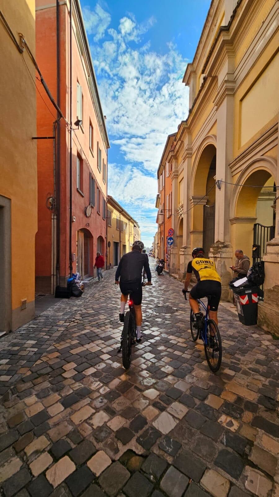 Dwaj kolarze jadący wąską, brukowaną uliczką włoskiego miasteczka między kolorowymi kamienicami w odcieniach pomarańczu i żółci pod błękitnym niebem.