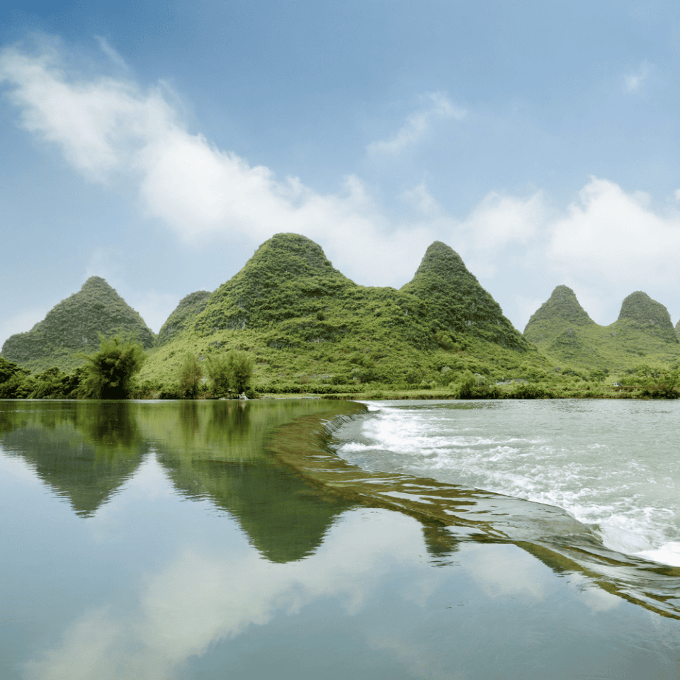 Zielone krasowe wzgórza odbijające się w spokojnej rzece Li w okolicach Yangshuo, pod błękitnym niebem z lekkim zachmurzeniem.